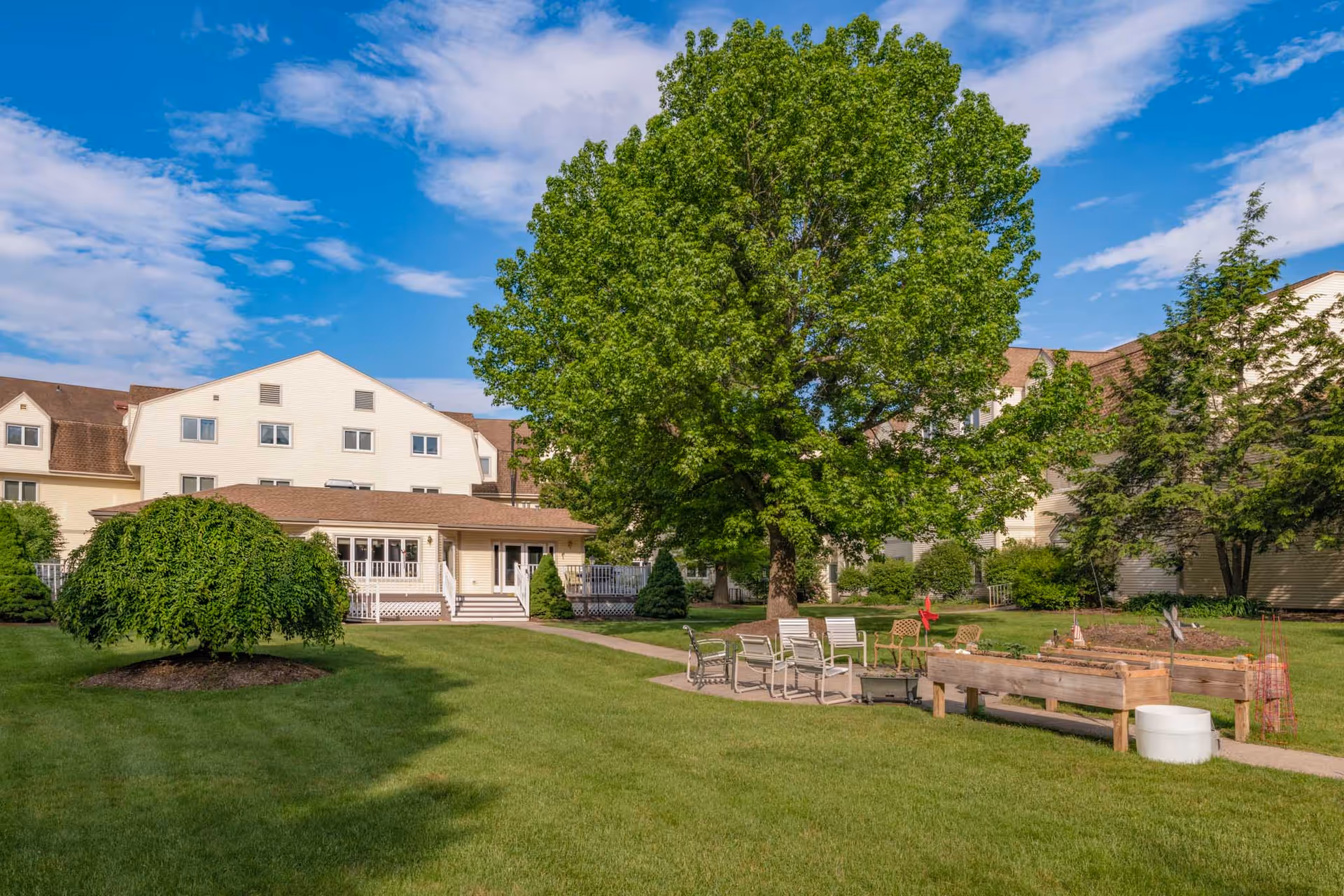 Outdoor view of Holiday Farmington facility showing a large green lawn with a variety of trees and shrubs. There is a paved walkway leading to a seating area with several chairs and a raised garden bed. The building is visible in the background under a partly cloudy blue sky.