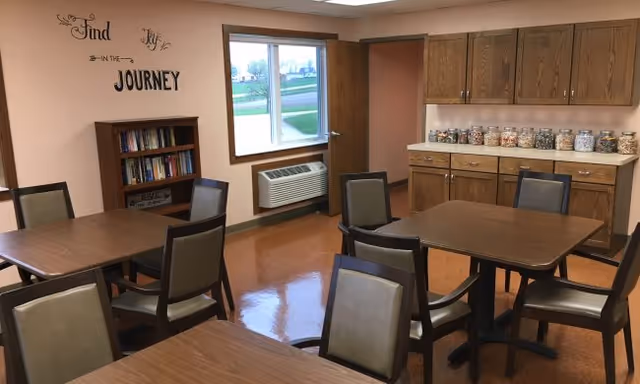 A cafe-style dining area with square tables, chairs, a cabinet with jars, and a window.