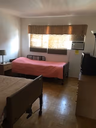 Small shared bedroom with two hospital-style beds, a window with a valance and an air conditioner, a dresser with a TV, and a nightstand.