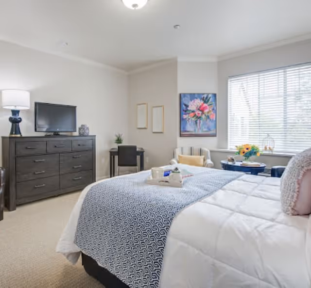 A bright and cozy bedroom with a large bed covered in white and blue bedding. There is a dark wooden dresser with a lamp and a TV on top, a small desk with a chair in the corner, and a comfortable armchair next to a window with blinds. The walls are decorated with framed artwork and a colorful floral painting.