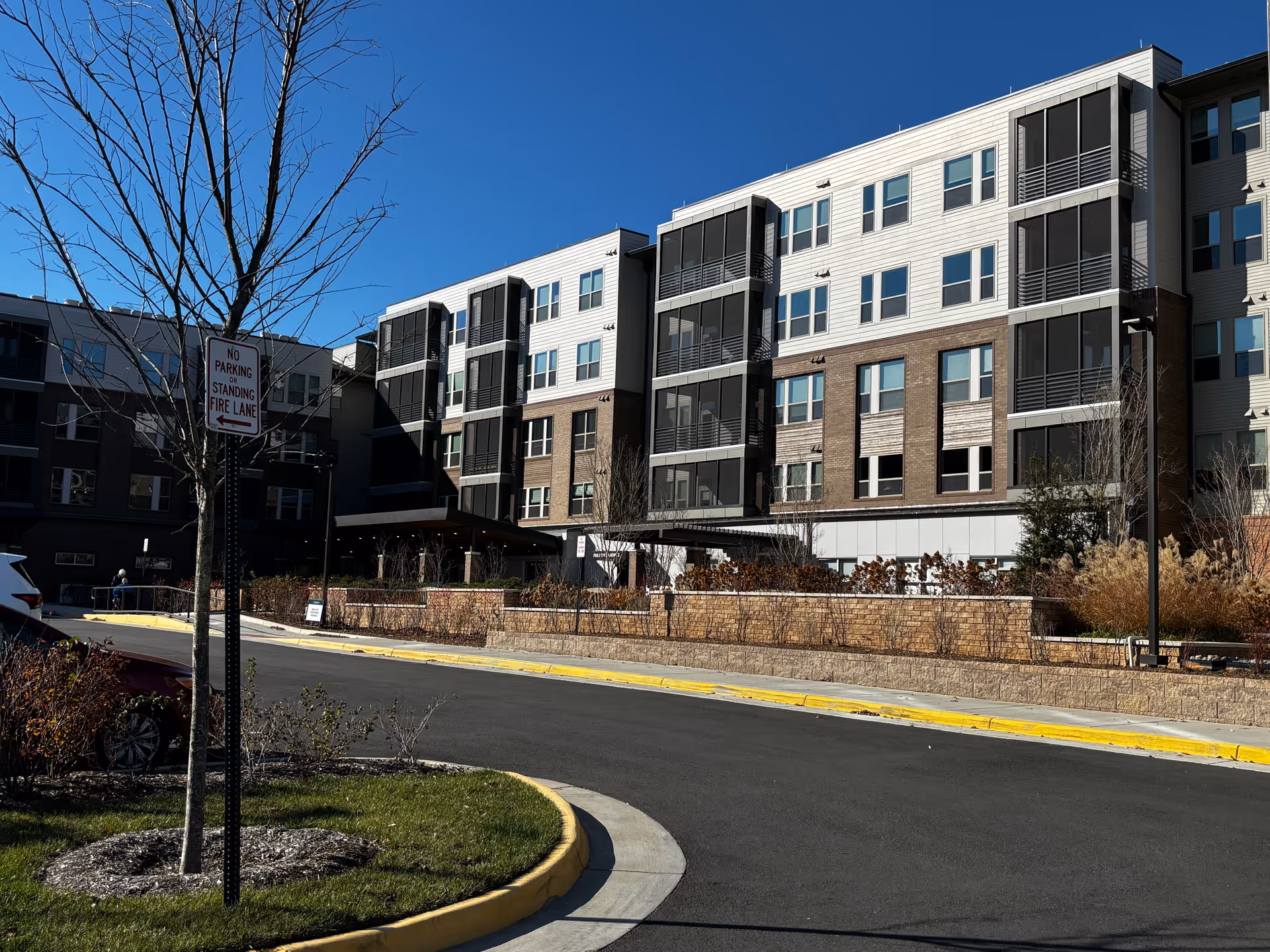 Exterior view of a modern multi-story senior living facility building under a clear blue sky. The building features large windows and balconies with black railings. In the foreground, there is a curved driveway with a 'No Parking or Standing Fire Lane' sign and some landscaping including a tree and bushes.