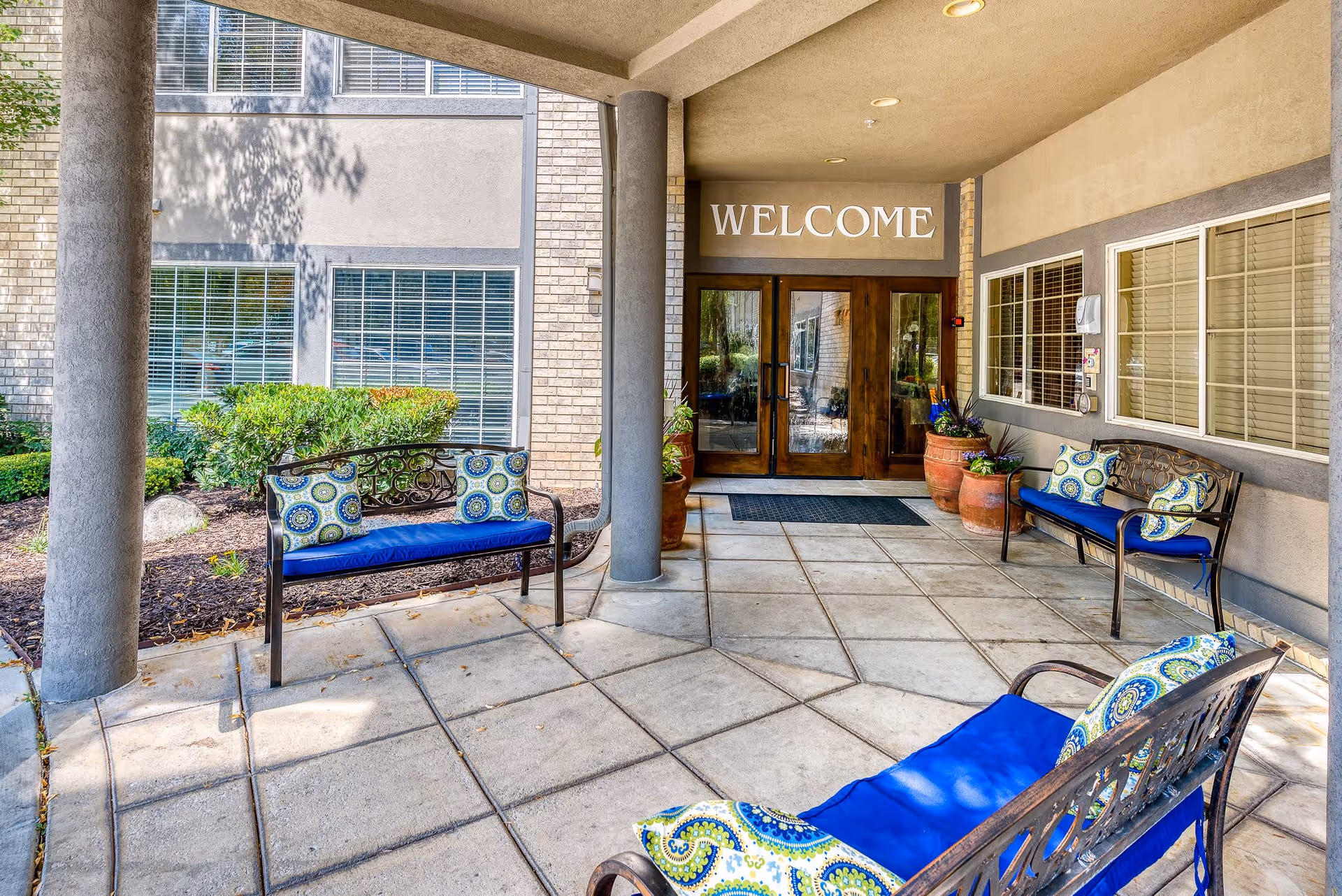Covered entrance area of a building with a 'WELCOME' sign above double wooden doors. There are three metal benches with blue cushions and patterned pillows arranged around the entrance. Large potted plants are placed near the doors, and windows with blinds are visible on the building walls.