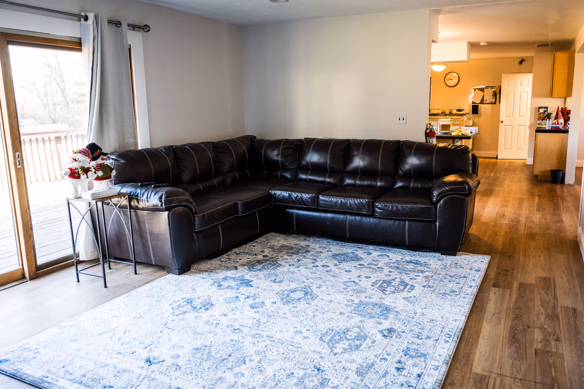 A living room with a large dark brown leather sectional sofa on a blue and white patterned rug. To the left, there is a sliding glass door with white curtains leading to an outdoor deck. A small side table next to the sofa holds a decorative snowman figure. The room has wooden flooring and leads into a kitchen area in the background.