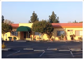 Exterior view of a single-story senior living facility building with a yellow facade, red tile roof, and a green awning over the entrance. There are trees and shrubs in front of the building and an empty parking lot in the foreground.