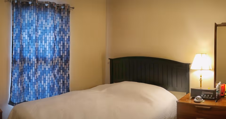 Bedroom with a made bed, dark wooden headboard, patterned blue curtains and a bedside lamp on a wooden dresser.