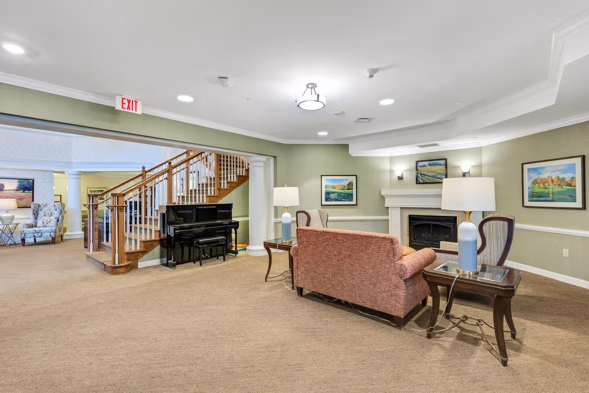 A spacious living room area in a senior living facility featuring a pink upholstered sofa, two wooden side tables with lamps, a fireplace with a mantle, framed landscape paintings on the walls, a wooden staircase with white railings, and a black upright piano. The room has beige carpeting, green walls with white trim, and recessed ceiling lights.
