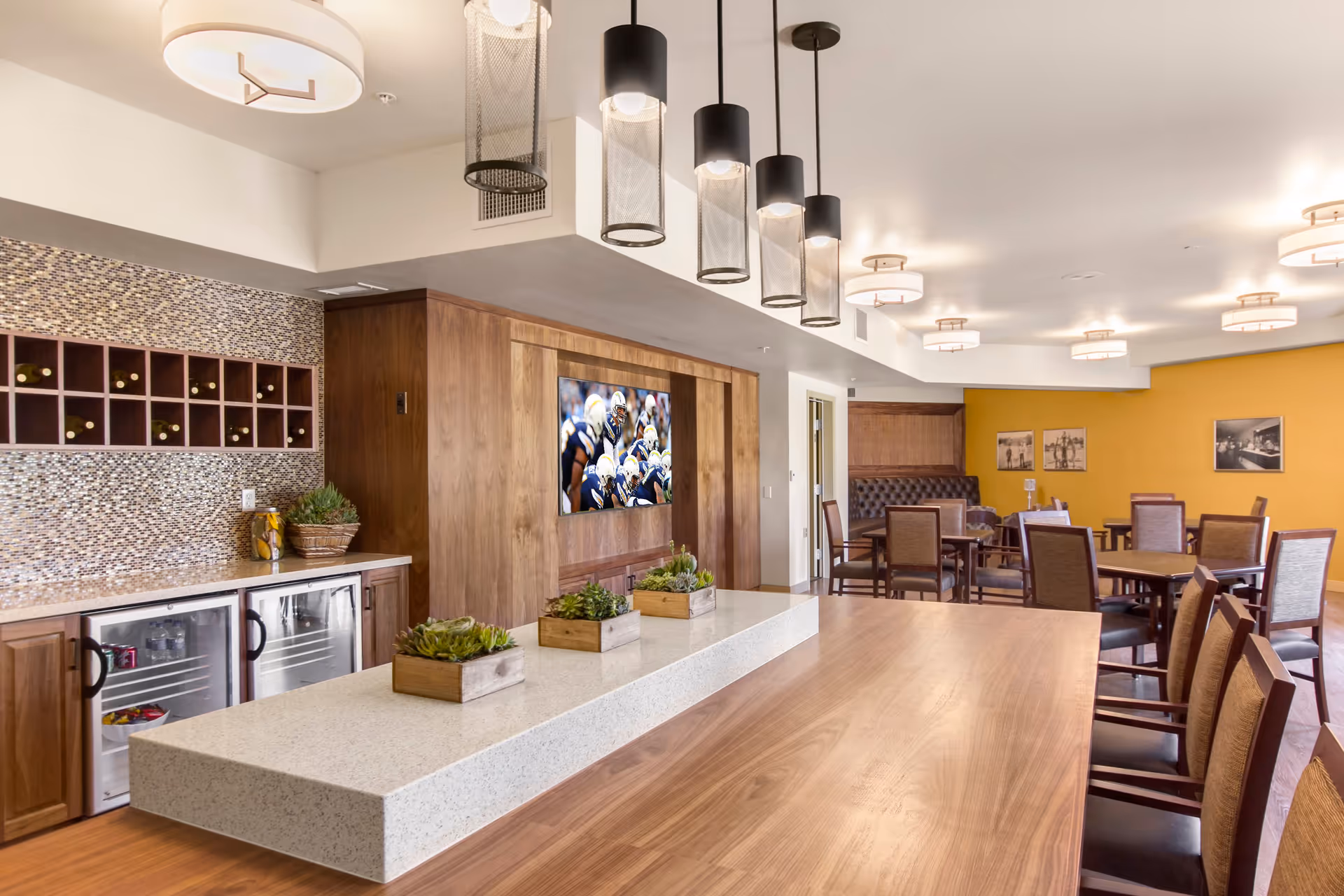 A modern dining room with wooden tables and chairs, a long countertop with small potted plants, pendant lights hanging from the ceiling, a wall-mounted TV showing a football game, and a wine rack above a countertop with mini refrigerators underneath. The walls are painted yellow and white with framed pictures on the yellow wall.