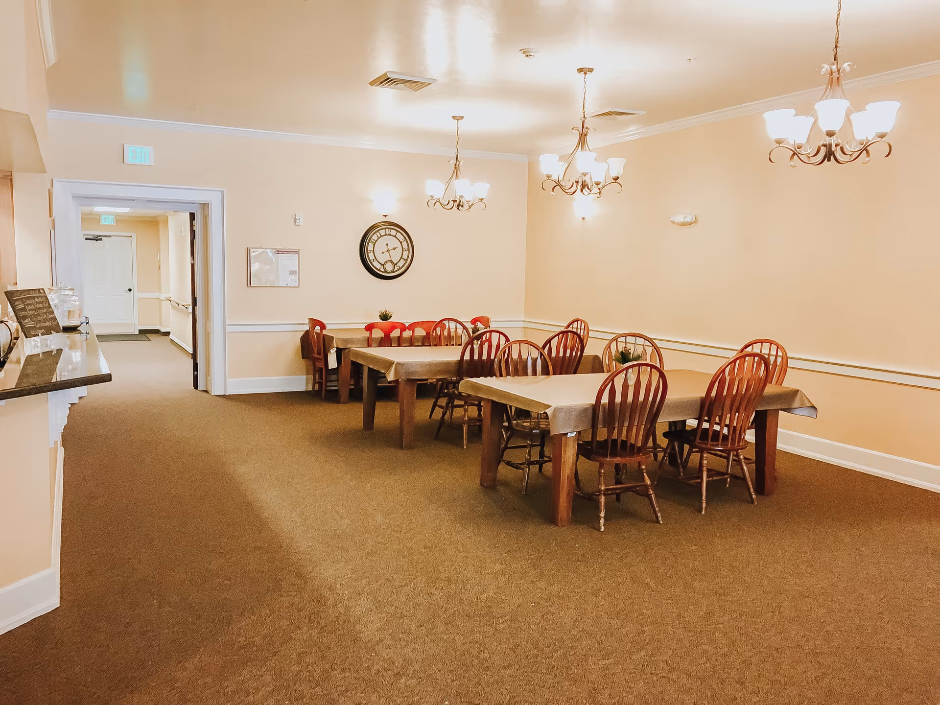 A dining room in an assisted living facility with two rectangular tables covered with beige tablecloths, surrounded by wooden chairs. The room has beige walls, carpeted floor, three chandeliers hanging from the ceiling, a large wall clock, and a hallway visible through an open doorway.