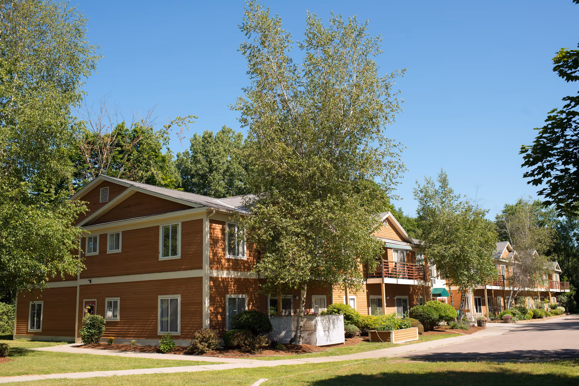 Exterior view of a two-story residential building with brown siding and white trim, surrounded by green trees and shrubs under a clear blue sky.