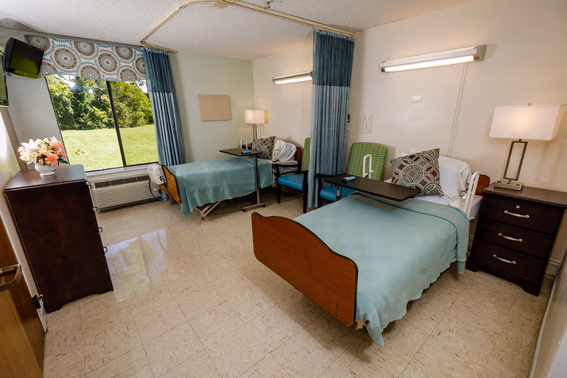 A bright and clean shared bedroom in a senior living facility with two hospital-style beds, each with a green blanket and patterned pillows. Each bed has an overbed table and a green chair beside it. There is a large window with blue curtains letting in natural light and showing a green outdoor area. The room also contains two dark wood dressers, two lamps, and a wall-mounted TV.