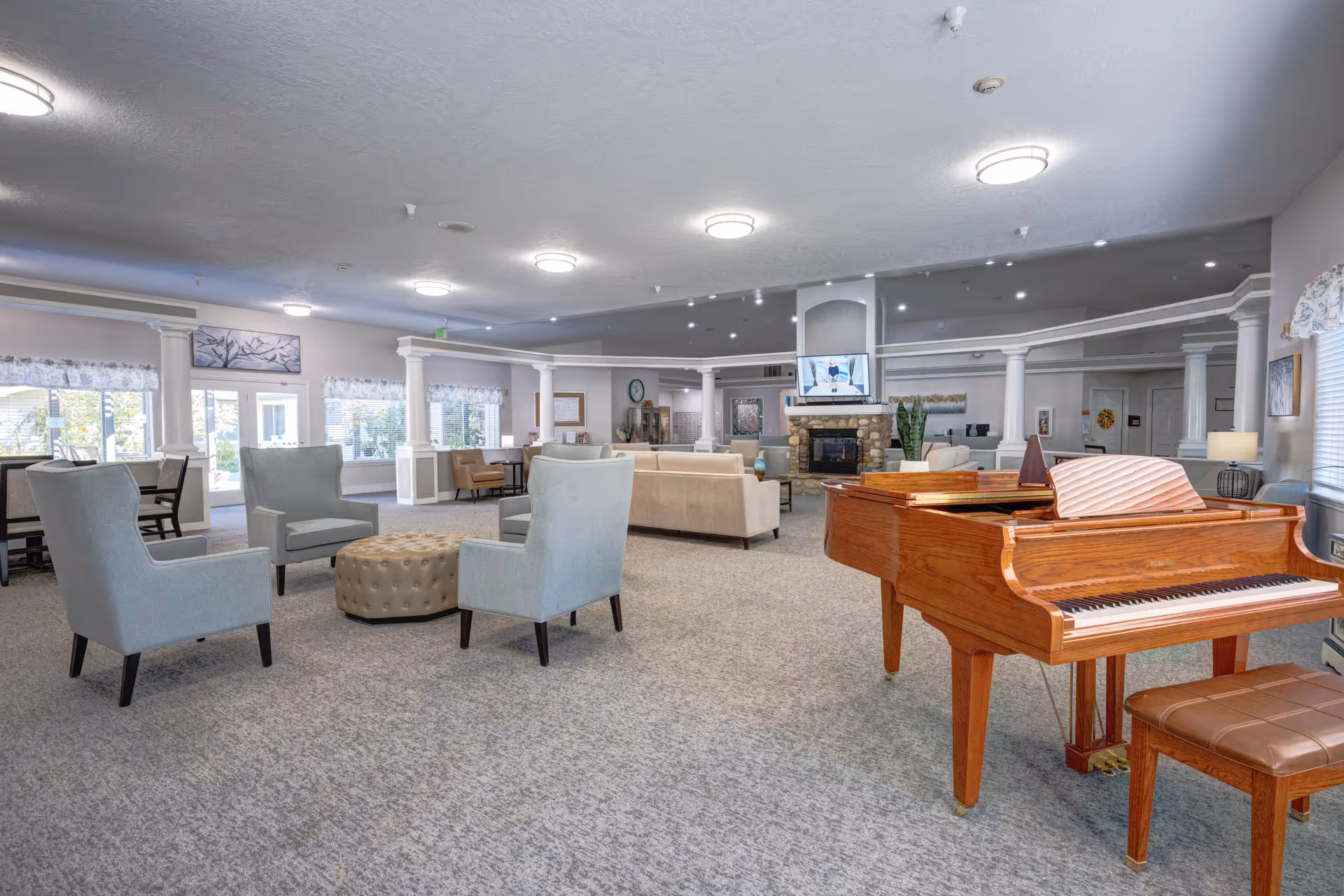 Spacious senior living common area with seating arrangements, a grand piano, and a fireplace with TV in the background.