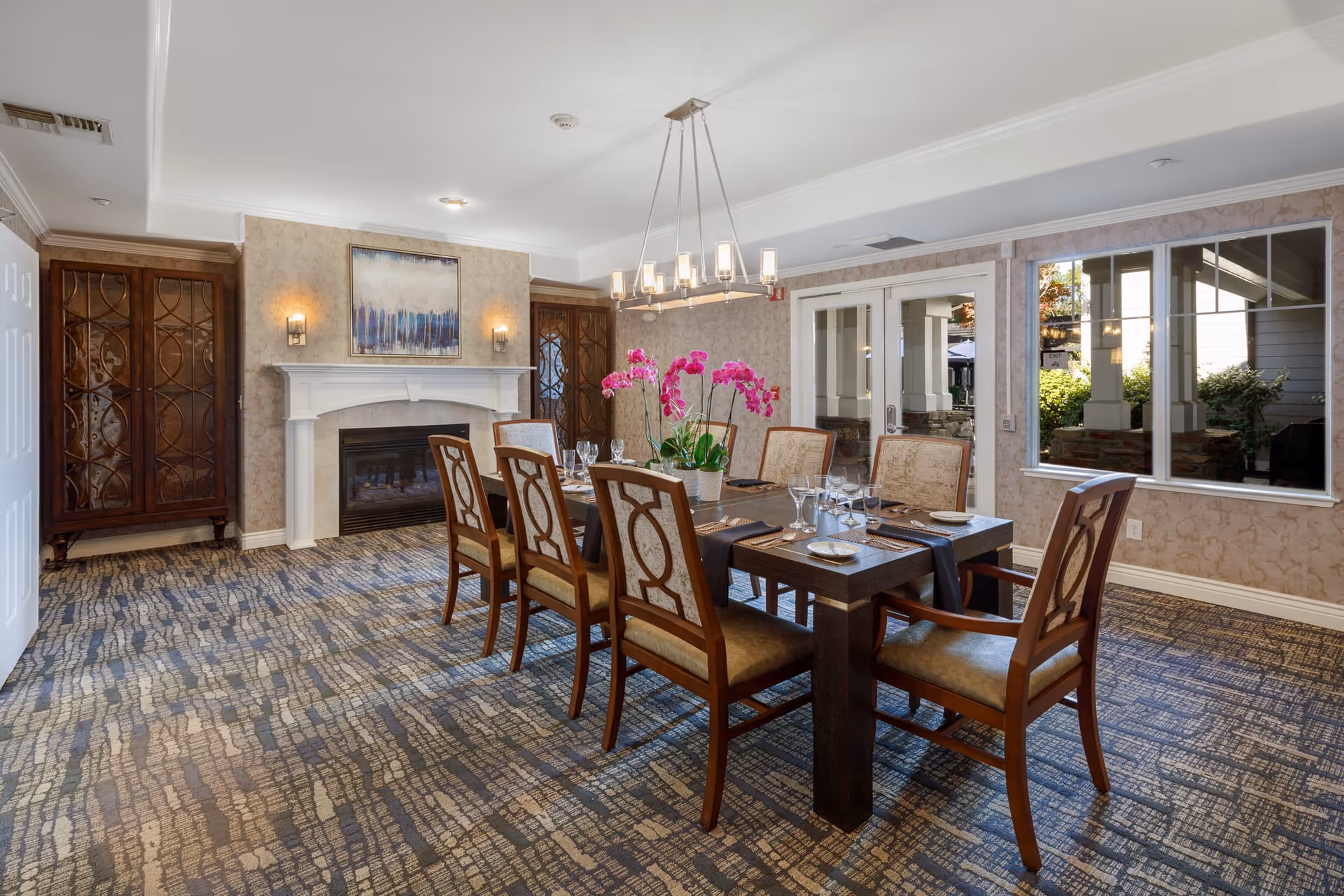 A well-lit dining room with a rectangular wooden dining table set for six people. The table has a centerpiece with pink orchids and is surrounded by six upholstered chairs with wooden frames. The room features a fireplace with a white mantel, two wooden cabinets with glass doors on either side, a modern chandelier above the table, patterned carpet flooring, and large windows and glass doors that look out to an outdoor area.