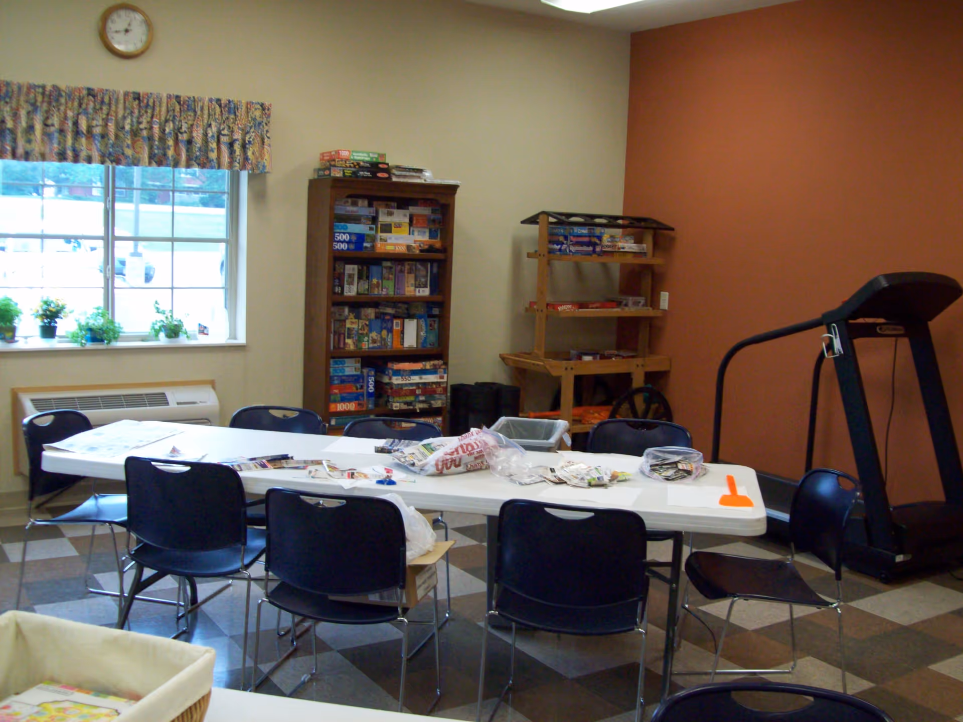 A senior living activity room featuring a long folding table with chairs, shelves of board games, and a treadmill by a window.