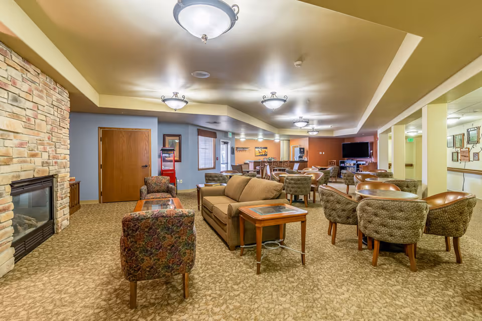 A spacious senior living facility common area with multiple seating arrangements including sofas, armchairs, and round tables with chairs. The room features a stone fireplace on the left, a flat-screen TV mounted on the far wall, and warm lighting from ceiling fixtures. The carpeted floor and neutral wall colors create a cozy atmosphere.