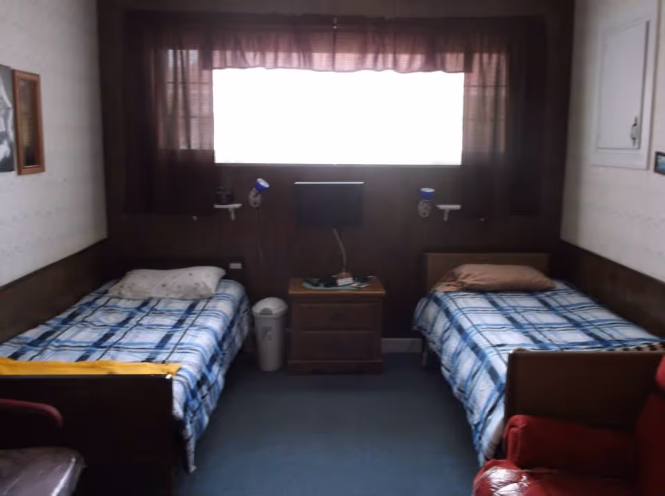 A small bedroom with two single beds placed parallel to each other against opposite walls. Each bed has a plaid blue and white blanket and a pillow. Between the beds is a wooden nightstand with a small TV mounted on the wall above it. There are two wall-mounted reading lamps above each bed. A window with dark curtains is centered on the back wall, letting in some light. The room has a carpeted floor and wood-paneled walls.