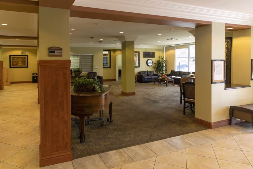 Spacious senior living facility lounge with seating, a piano, columns and tiled floor.