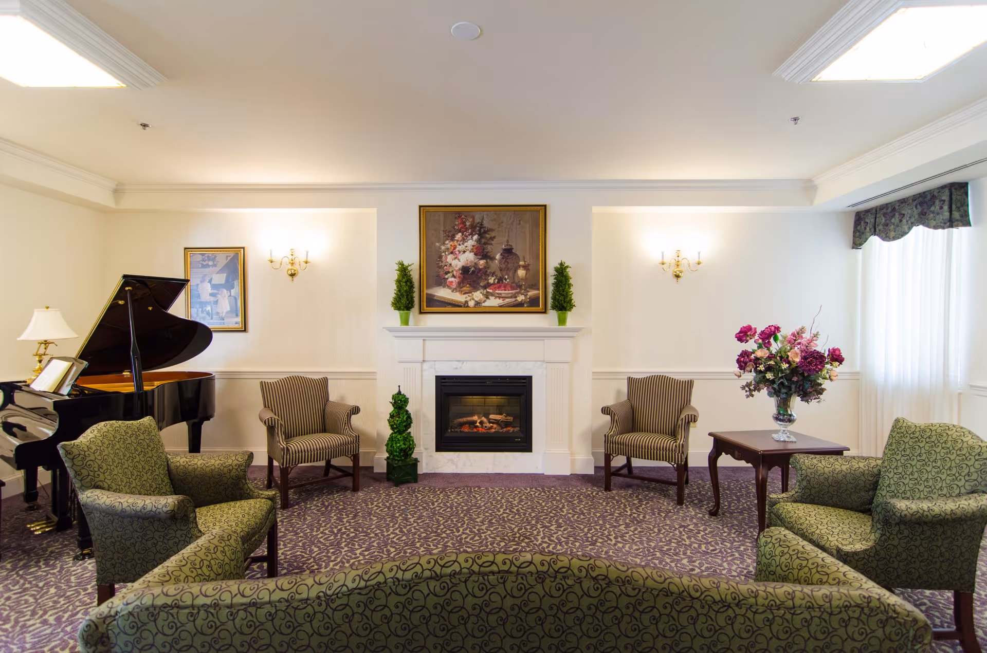 A cozy living room area with patterned green armchairs and a sofa arranged around a white fireplace with a floral painting above it. There is a grand piano on the left side, two striped armchairs near the fireplace, a small table with a vase of flowers on the right, and soft lighting from wall sconces and ceiling lights.