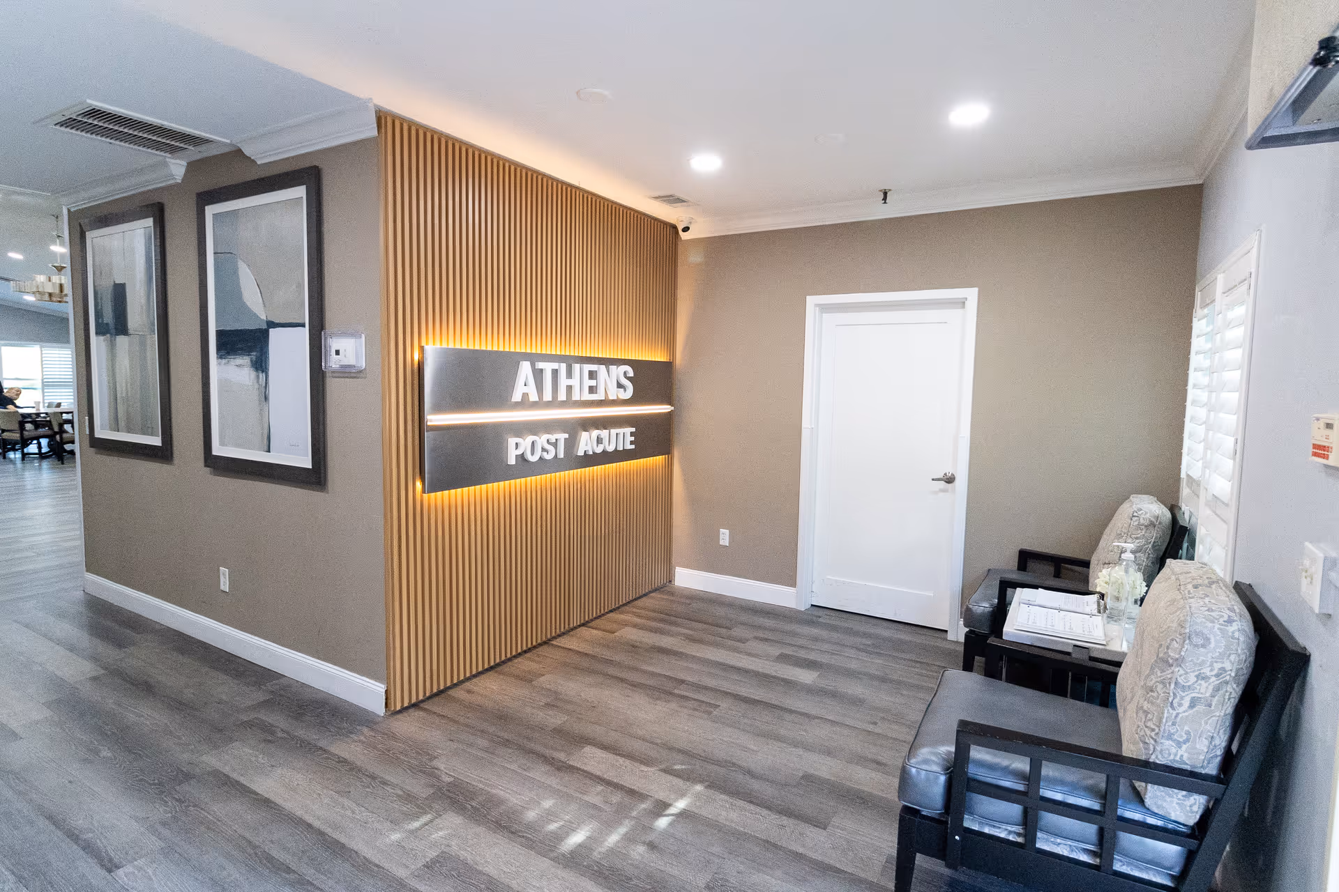 Interior view of a hallway in a senior living facility with a wooden slat wall featuring illuminated signage that reads 'ATHENS POST ACUTE'. The hallway has gray wood flooring, beige walls, two framed abstract artworks, a white door, and two cushioned chairs with a small table between them.