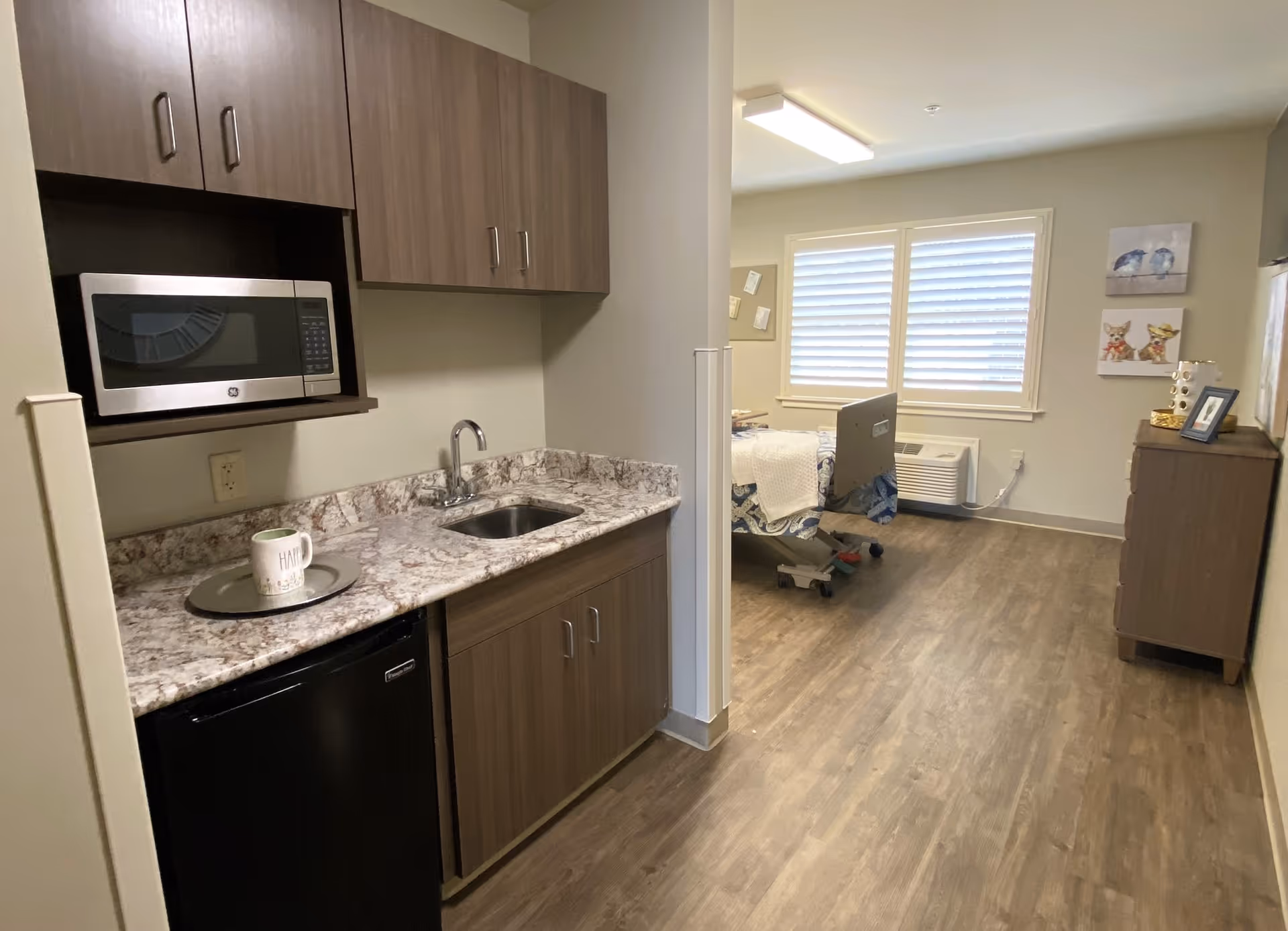 A small kitchenette area with a microwave, sink, and mini refrigerator on the left side, leading into a room with a hospital bed, dresser, and two windows with blinds. The floor is wood-style laminate, and the walls are light-colored.