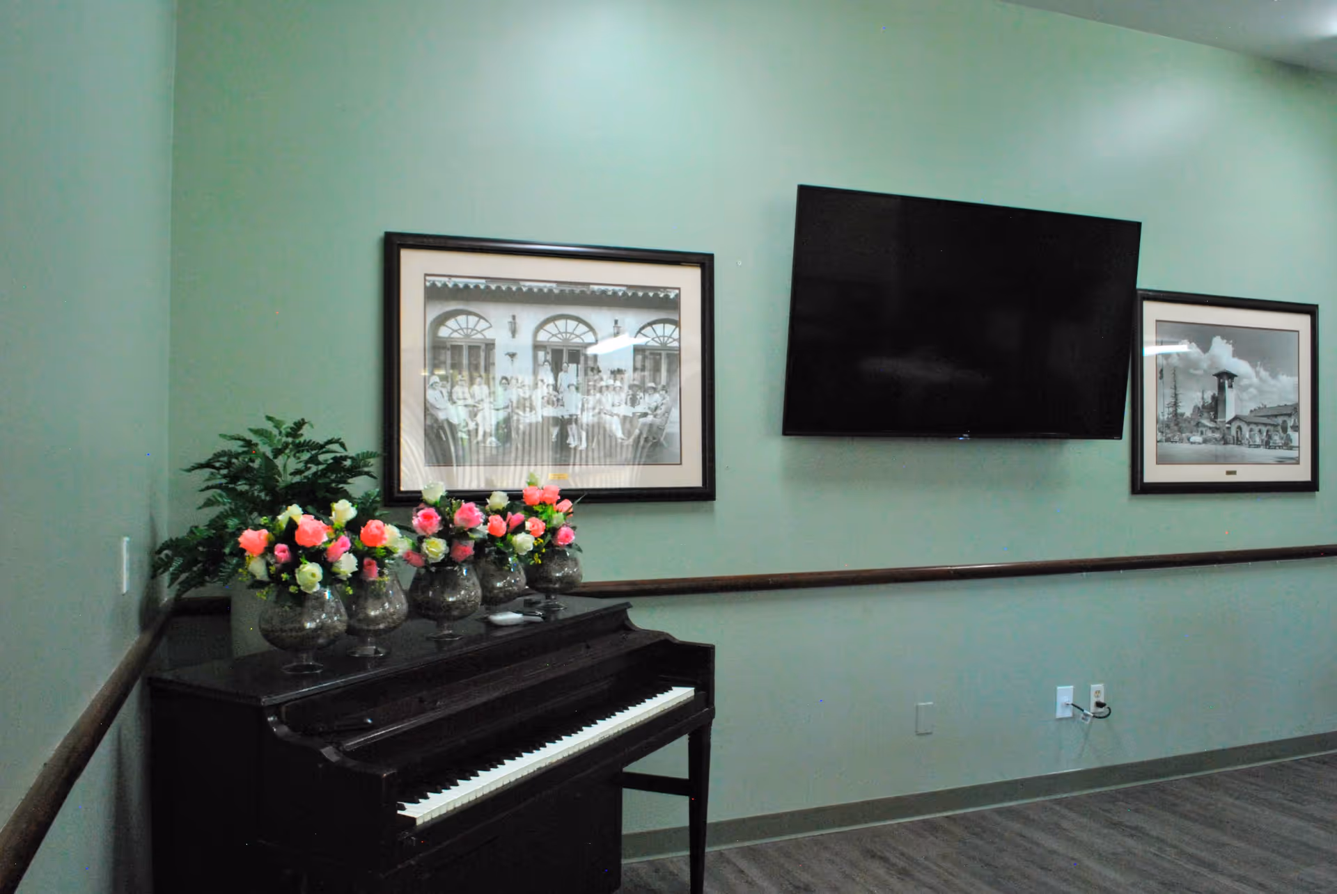 Interior room with a black upright piano adorned with five glass vases filled with pink and white flowers. Above the piano are two framed black and white photographs on a light green wall. A flat-screen TV is mounted on the wall between the pictures. The floor is wood, and there is a wooden handrail along the wall.