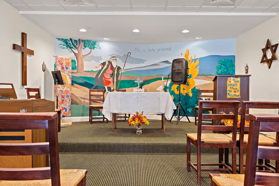 Interior of a small chapel or worship room with wooden chairs arranged facing a table covered with a white cloth. Behind the table is a mural depicting a biblical scene with a man holding a staff near a burning bush and the words 'This is holy ground'. There is a wooden cross on the left wall and a Star of David on the right wall. A lectern with a sign that reads 'It's no small thing to believe' is on the right side of the room.