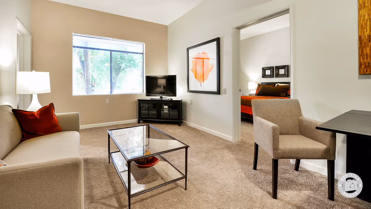 A cozy living room with beige carpet and walls, featuring a beige sofa with a red pillow, a glass coffee table, a beige armchair, a black TV stand with a flat-screen TV, and a large window with blinds. A doorway leads to a bedroom with a bed that has red and brown bedding. There is framed artwork on the walls and a table next to the armchair.