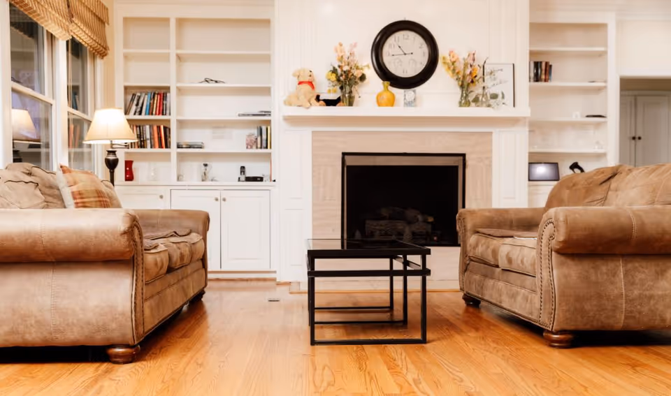 A cozy living room with two brown leather sofas facing each other, a black nested coffee table in the center, a fireplace with a clock and decorative items on the mantel, built-in white shelves with books and decor, and a floor lamp next to a window with wooden blinds.