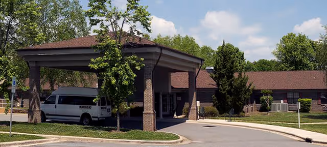 Front exterior view of UNC Rockingham Rehabilitation and Nursing Care Center showing a covered entrance with a white van parked underneath, surrounded by trees and greenery under a partly cloudy sky.