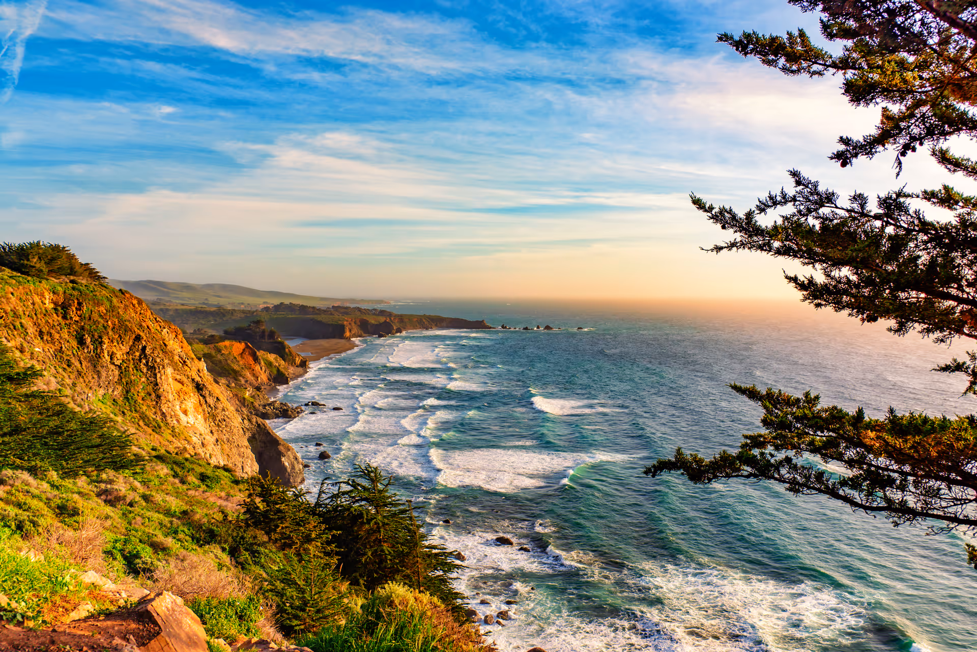 Scenic view of a coastal cliff with green vegetation overlooking the ocean with waves crashing against the shore under a partly cloudy sky during sunset.