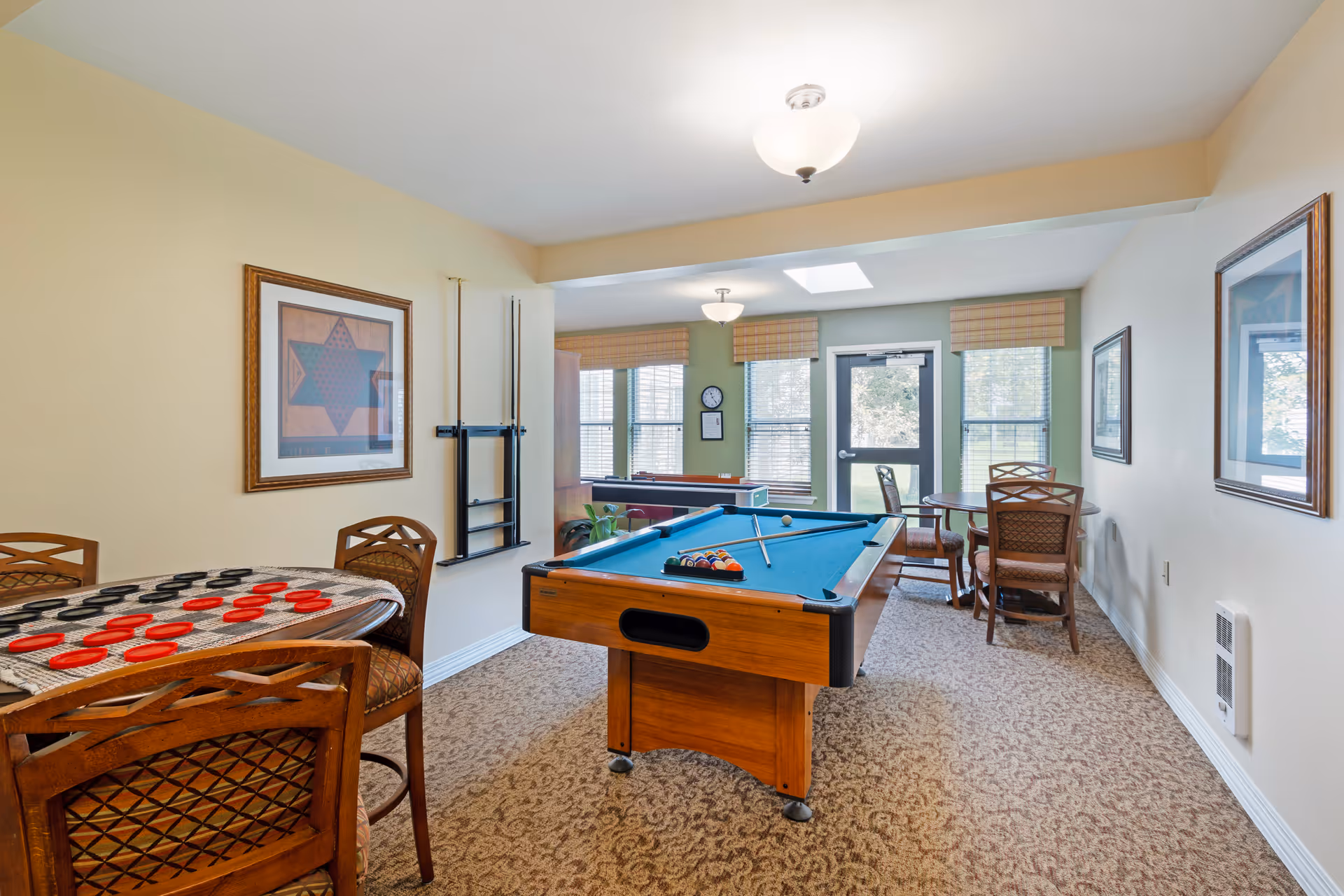 A recreational room with a pool table in the center, a round table with a checkers game on the left, and another round table with chairs near the windows. The room has beige walls, carpeted floor, framed artwork on the walls, and multiple windows letting in natural light.