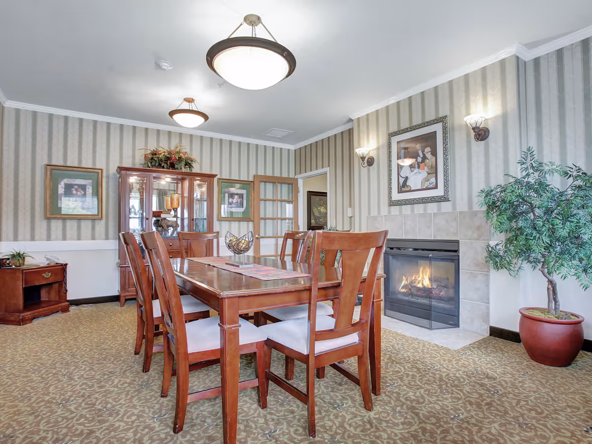 A dining room with a wooden table and chairs, a lit fireplace, china cabinet, framed artwork, and a potted plant.