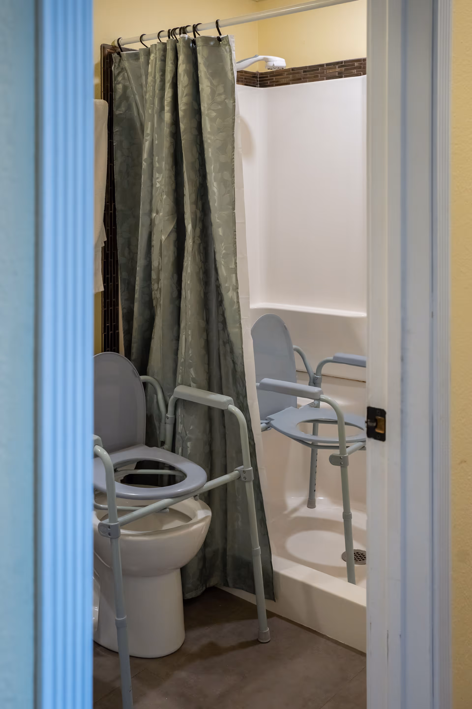 Small bathroom with a shower and two gray raised commode/shower chairs and a green shower curtain.