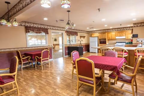 A dining area in a senior living facility featuring wooden floors, tables covered with red tablecloths, and wooden chairs with red cushions. The room includes a kitchen area with wooden cabinets, a white refrigerator, and a stove. Decorative light fixtures hang from the ceiling, and windows with valances allow natural light into the space.
