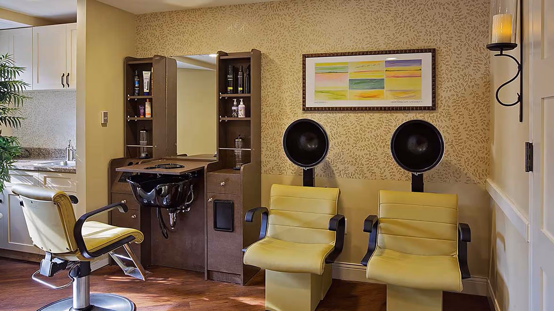 Interior of a salon area in a senior living facility with two yellow salon chairs in front of hair drying stations, a styling chair, a sink, and a cabinet with hair care products. The walls have a patterned wallpaper and a framed abstract artwork is hung above the drying stations.