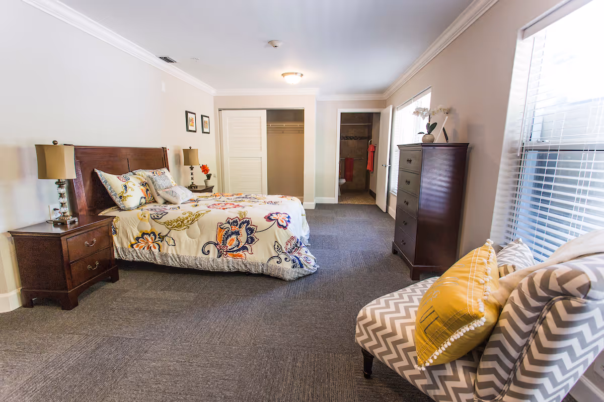 A spacious bedroom with a large bed featuring floral bedding, two bedside tables with lamps, a wooden dresser, and a patterned armchair with a yellow pillow. The room has carpeted flooring, two windows with blinds, and an open door leading to a bathroom.