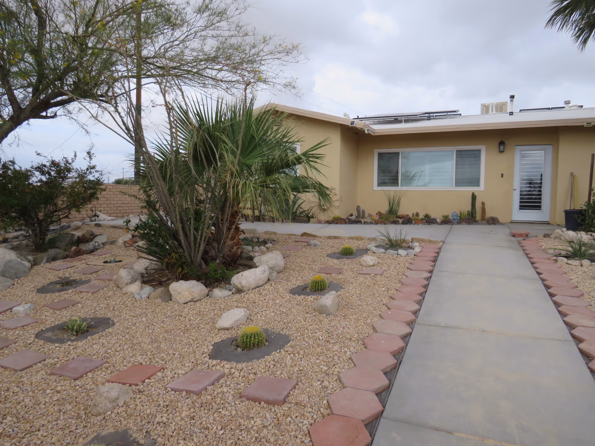 Front yard of a single-story building with a beige exterior, featuring a concrete walkway bordered by red hexagonal and square pavers leading to a white door with a window. The yard is landscaped with gravel, rocks, small cacti, and various desert plants including a palm tree and other shrubs. The sky is cloudy.