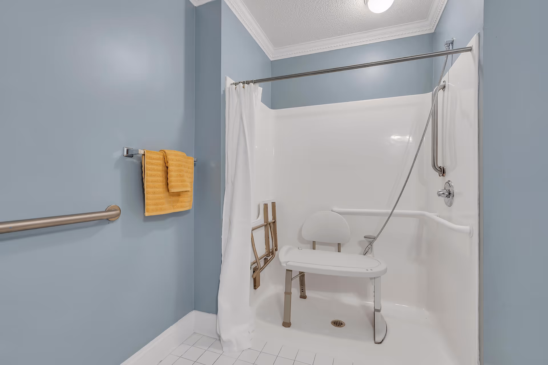 Accessible shower area with a white shower chair, grab bars, and a handheld showerhead. The walls are painted blue, and there is a white shower curtain partially drawn. A yellow towel hangs on a towel rack on the left wall.