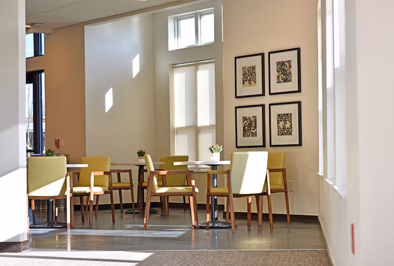 Sunlit dining area with round tables, green upholstered chairs, and framed artwork on the wall.
