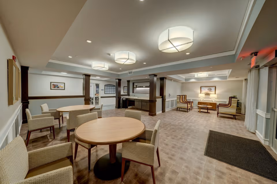 A spacious common area in a senior living facility featuring round wooden tables with cushioned chairs, a seating area with armchairs and a small table, soft lighting from ceiling fixtures, and neutral-toned walls and flooring.