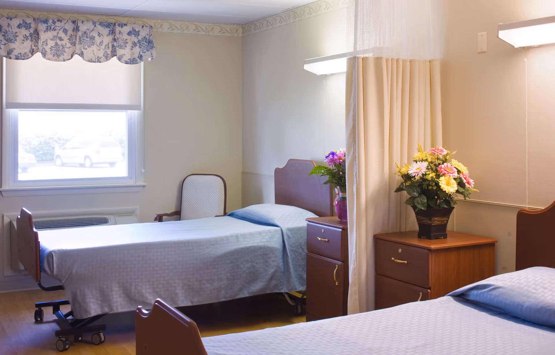 A bright room with two hospital-style beds separated by a privacy curtain. Each bed has light blue bedding. There are wooden nightstands next to each bed, one with a vase of colorful flowers. A window with a floral valance and a chair are visible in the background.