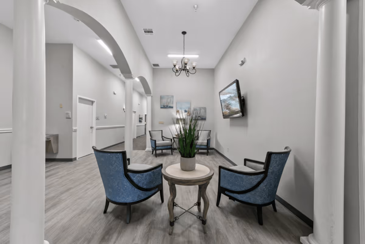A modern common area in a senior living facility featuring four blue upholstered chairs arranged around a small round wooden table with a potted plant. The room has light gray walls, wood flooring, white columns, a chandelier, a wall-mounted TV, and framed artwork on the far wall.