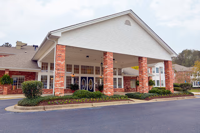 Front exterior view of a senior living facility with a large covered entrance supported by brick columns, landscaped bushes and flowers, and a paved driveway.