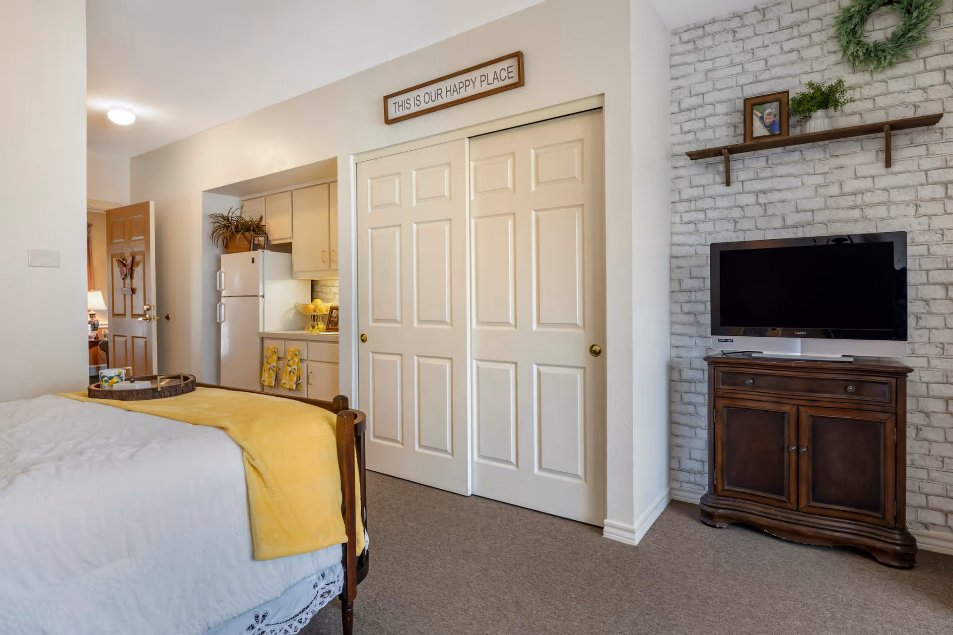 A cozy bedroom with a bed covered in white bedding and a yellow blanket. A wooden tray with a cup and a small plant sits on the bed. To the right, there is a wooden cabinet with a flat-screen TV on top, against a white brick accent wall with a shelf holding a framed photo and a green wreath. In the background, a small kitchen area with white cabinets, a refrigerator, and a countertop is visible. Above the closet doors, a sign reads 'THIS IS OUR HAPPY PLACE'.