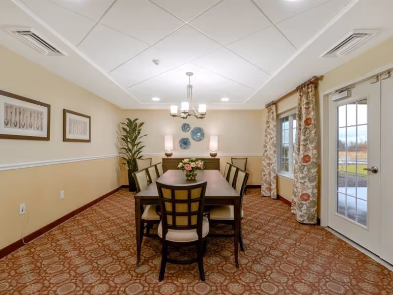 A dining room with a rectangular wooden table surrounded by eight chairs with cushioned seats. A vase with flowers is placed in the center of the table. The room has patterned carpet flooring, beige walls with white trim, and two framed pictures on the left wall. A potted plant is in the corner, and a sideboard with two lamps and decorative plates on the wall is at the far end. There is a window with floral curtains and a glass door on the right side, allowing natural light to enter.