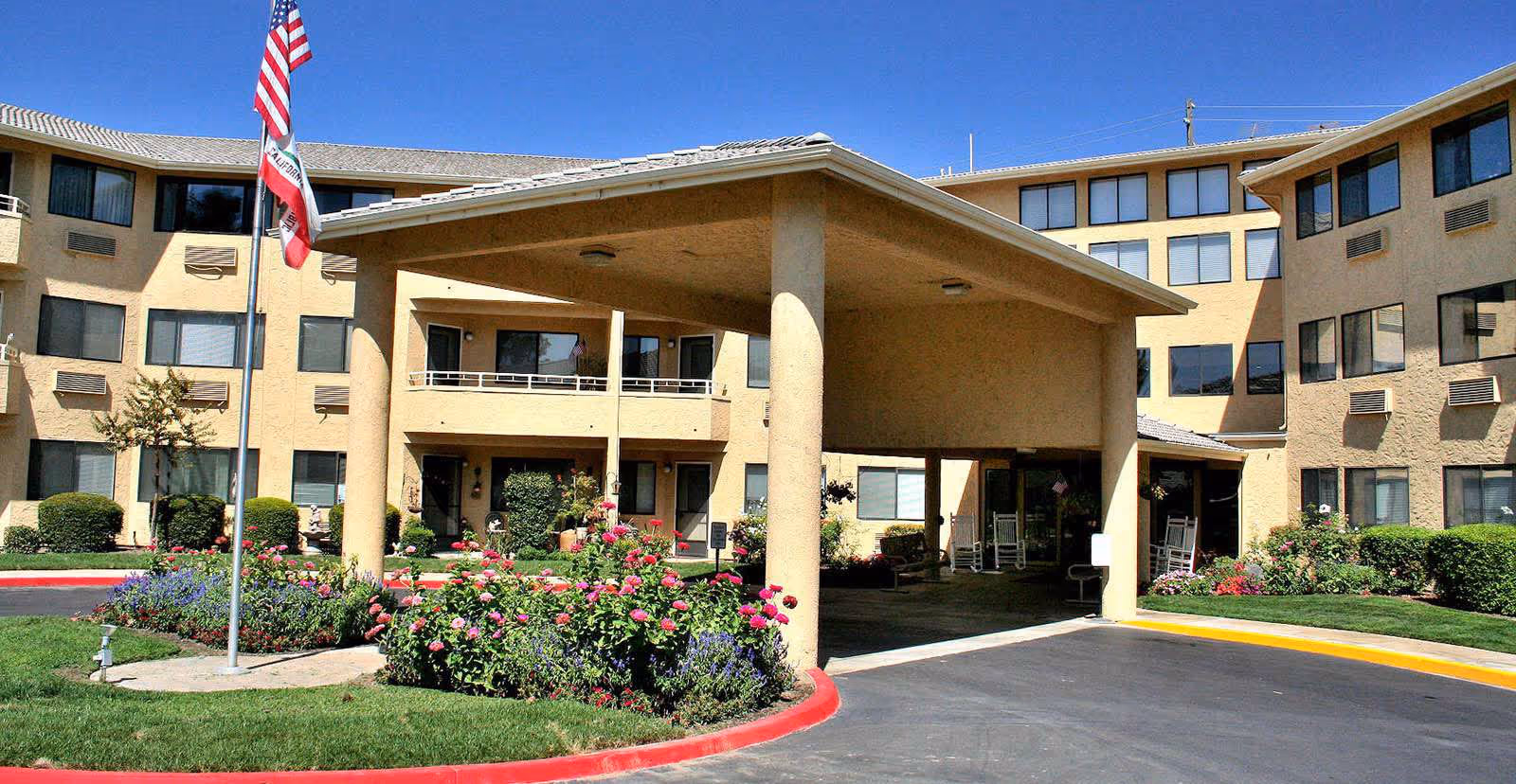 Exterior view of Walnut Park - A Provincial Senior Living Community, showing a multi-story beige building with numerous windows, a covered entrance supported by large columns, landscaped flower beds with pink and purple flowers, and an American flag and California state flag on flagpoles.