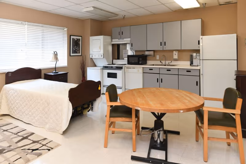 A room with a single bed covered with a white quilt, a wooden nightstand with a lamp, a round wooden table with two chairs, and a kitchenette area with cabinets, a stove, microwave, dishwasher, refrigerator, and a stacked washer and dryer unit. The walls are beige and there is a window with blinds letting in natural light.