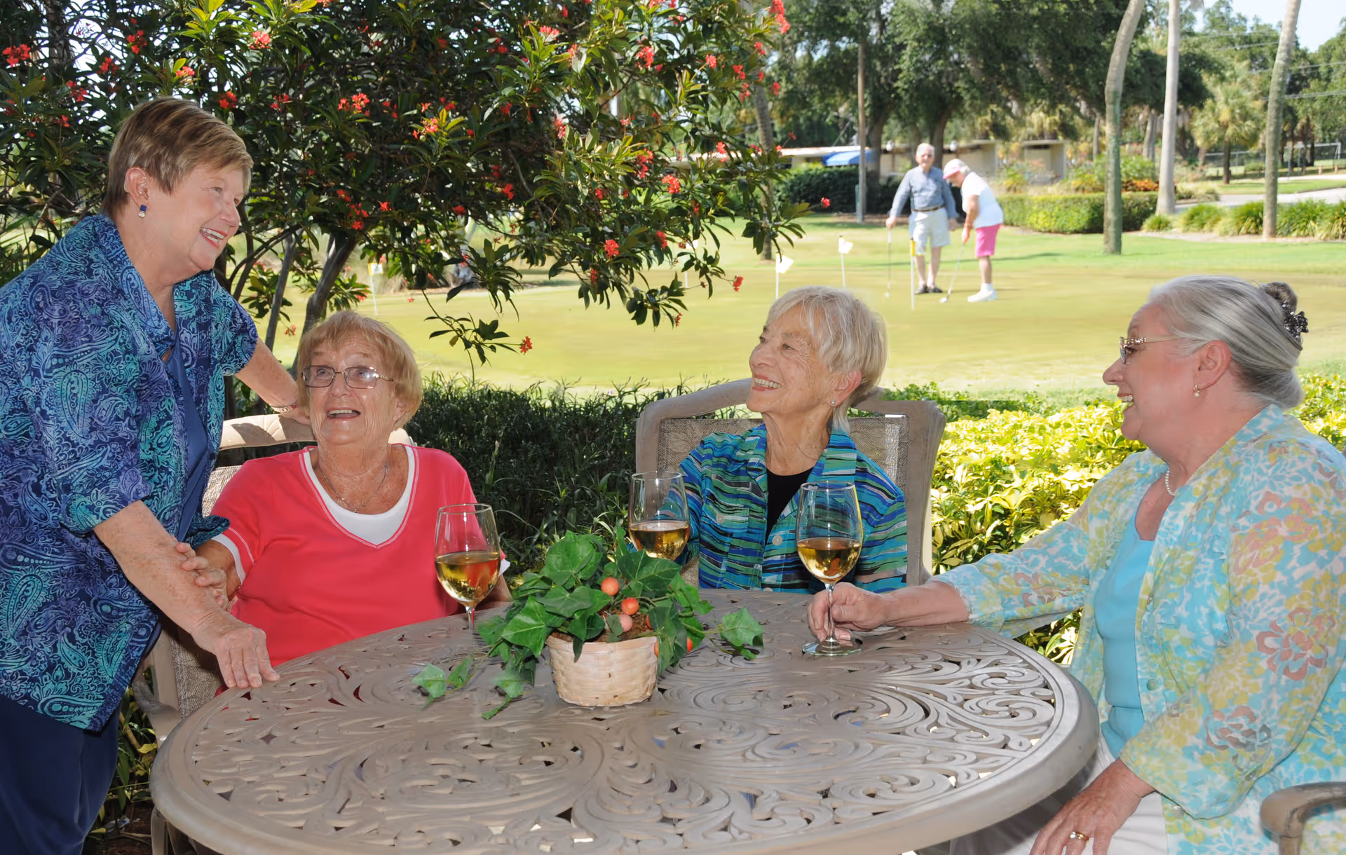 Four elderly women socialize at a round outdoor patio table with glasses of wine, with a golf green and players visible in the background.