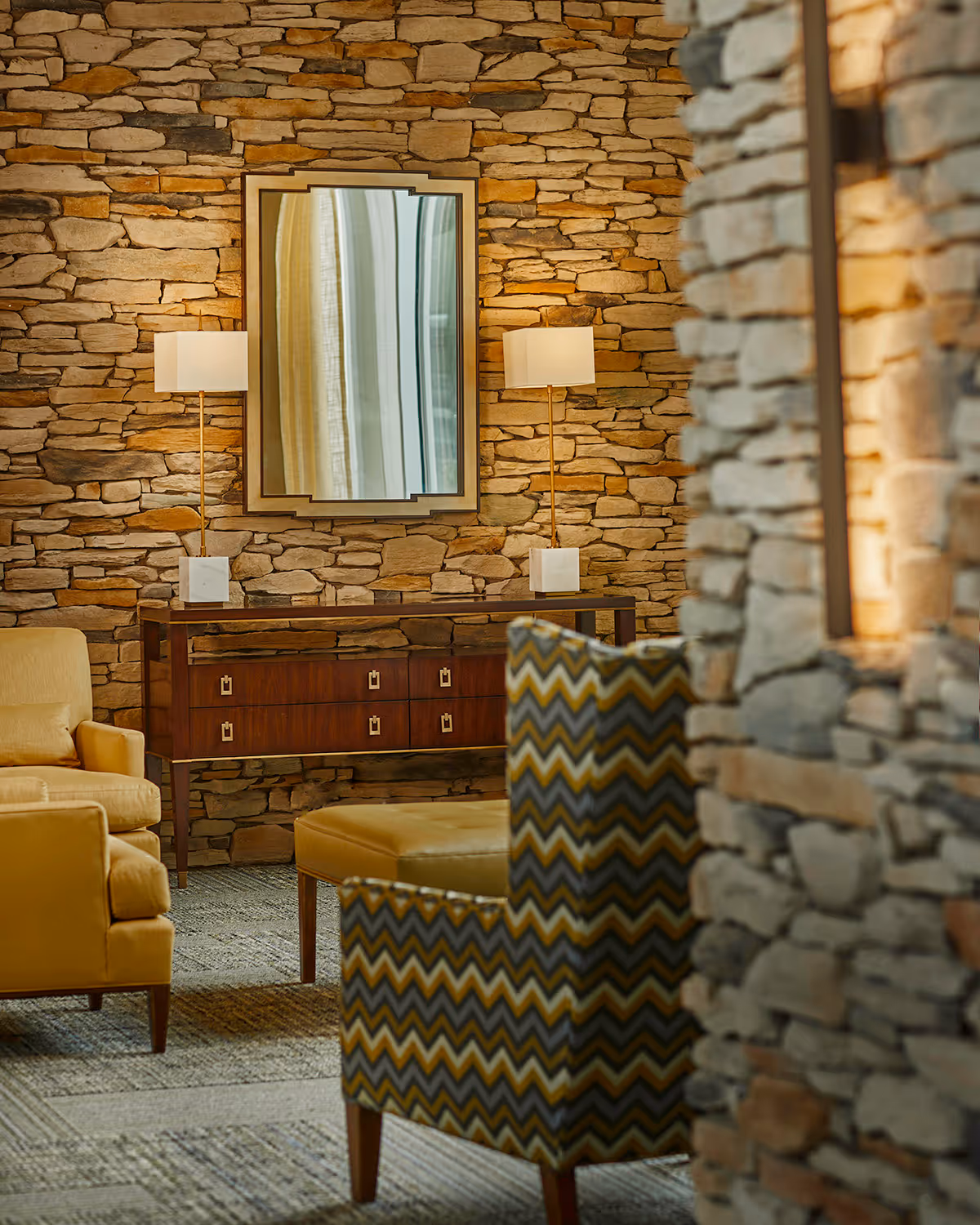 Cozy sitting area featuring a stone accent wall with a mirror and console table flanked by two lamps, yellow upholstered chairs, an ottoman, and a patterned armchair.