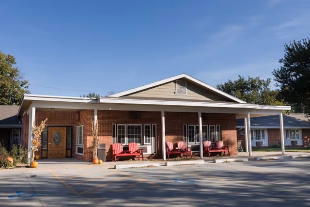 Exterior view of a single-story brick building with a covered porch featuring several red benches. The building has a wooden front door with glass panels and is surrounded by a paved parking area with marked spaces. Trees and clear blue sky are visible in the background.
