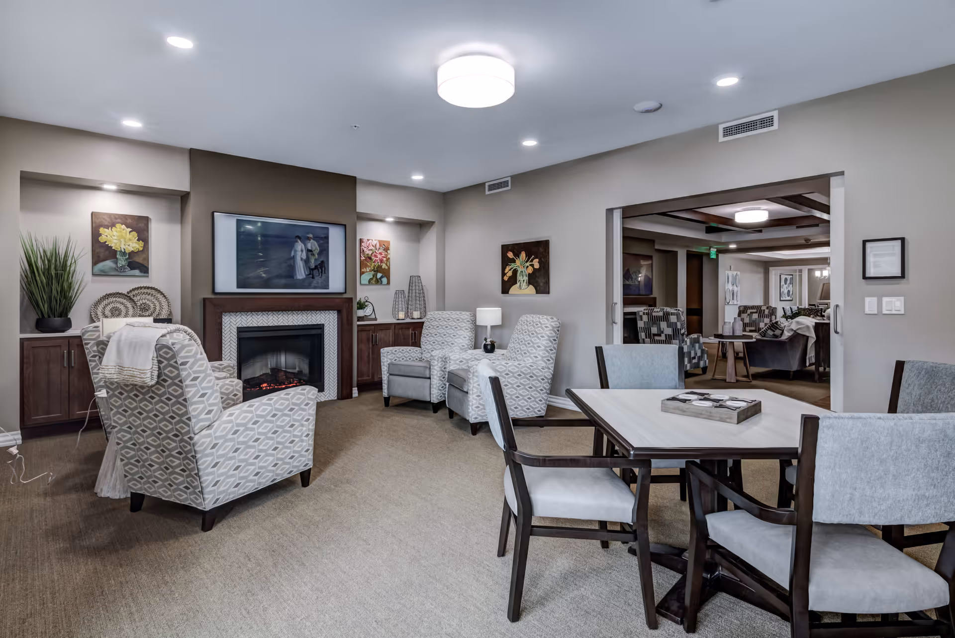 A cozy assisted living common area featuring a fireplace with a painting above it, surrounded by four patterned armchairs. There are built-in cabinets with decorative items and paintings on the walls. A table with four chairs is in the foreground, and another seating area is visible through a wide doorway. The room is softly lit with ceiling lights and a central light fixture.